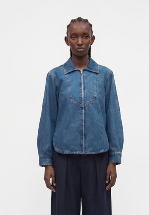 Mujer con cabello trenzado que lleva una chaqueta vaquera azul con cremallera y pantalones anchos oscuros, sobre un fondo blanco liso.