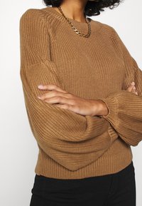 Överdimensionerad brun stickad tröja med ribbade ärmslut, texturerad tyg och bred rund halsringning. Matchas med ett guldkedjehalsband.