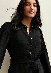 Vestido negro de botones con una tela texturizada, mangas abullonadas, cuello y cintura ceñida con cinturón. Presenta botones metálicos y líneas suaves.