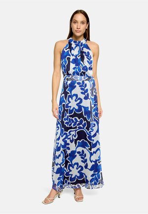 Kobieta ubrana w długą sukienkę bez rękawów z dekoltem halter, w niebiesko-biały kwiatowy wzór i beżowe sandały na obcasie na jednolitym tle.