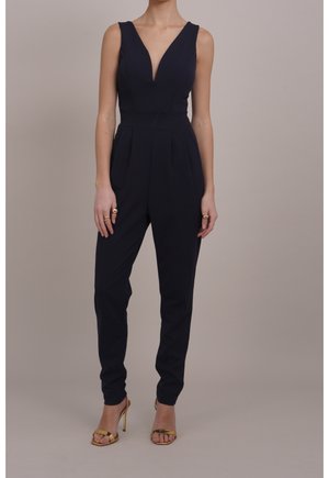 Marineblaue Overall mit tiefem V-Ausschnitt, ärmellosem Design und zulaufenden Beinen. Verfügt über eine plissierte Vorderseite und eine taillierte Passform, kombiniert mit goldenen Sandalen mit hohen Absätzen.