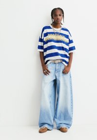 Gestreiftes, blau-weißes Oversized-T-Shirt mit gelbem Text, kombiniert mit hellblauen, weit geschnittenen Jeans und braunen Sneakers.