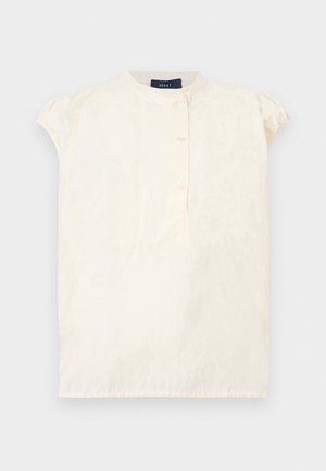 Kvinde iført en ærmeløs cremefarvet bluse, sort-hvide knælange shorts med nålestriber og sorte klipklapper, stående med den ene hånd i lommen.