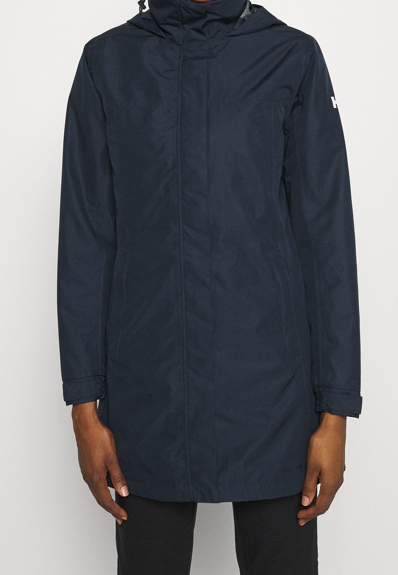 Personne portant un long manteau à capuche bleu marine avec des poches avant, debout devant un fond clair uni.