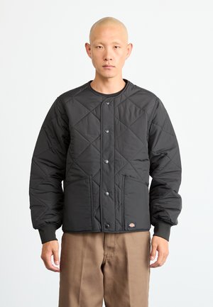 Man with shaved head wearing a black quilted jacket with snap buttons and brown pants, standing against a plain white background.
