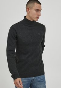 Pulover turtleneck gri închis, cu textură tricotată, croială slim, cu manșete și tiv ribbate, având un detaliu mic cu logo pe piept. Asortat cu blugi albaștri.