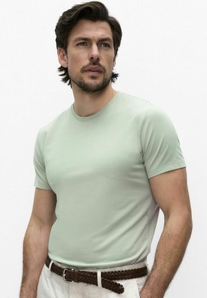 Man met donker haar en baard, draagt een lichtgroen aansluitend T-shirt, witte broek en gevlochten bruine riem, iets naar links gericht tegen een effen achtergrond.