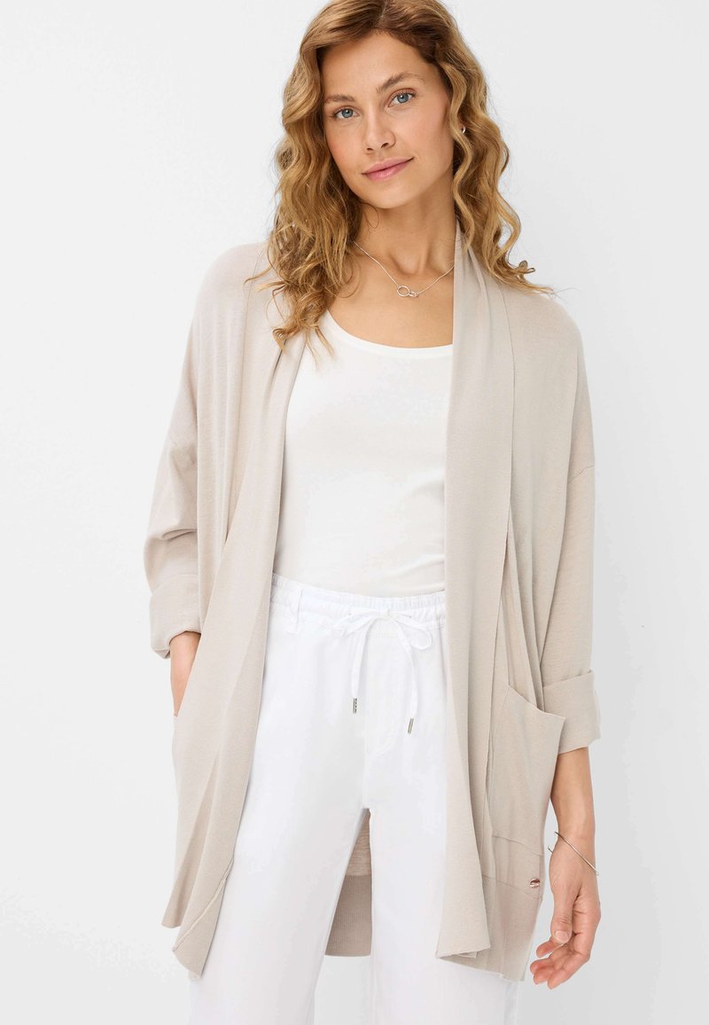 Cardigan beige à ouverture devant avec une coupe décontractée, des manches longues et des poches, associé à un haut blanc et un pantalon blanc à cordon de serrage. Texture lisse.