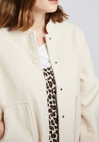 Veste en polaire crème avec un col haut, des fermetures à pression et deux poches frontales. Portée par-dessus une robe à imprimé léopard. Texture douce, coupe décontractée.