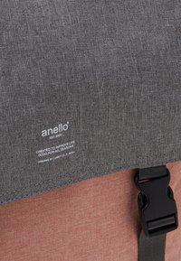 Bolsa mensajera de tela gris con una solapa gris más oscuro, que presenta una hebilla negra, una sección inferior naranja y una etiqueta de marca en texto blanco.