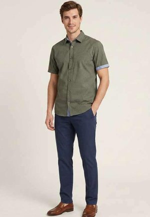 Homme debout portant une chemise verte à manches courtes boutonnée, un pantalon bleu marine et des chaussures en cuir marron, sur un fond clair uni.