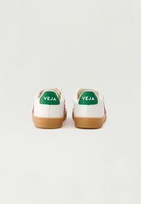 Baskets blanches en cuir avec des accents rouges, des languettes de talon vertes portant le logo "VEJA" et des semelles en caoutchouc naturel ; design minimaliste et lacets plats.