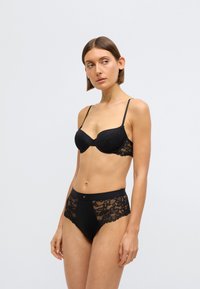 Čierna čipkovaná podprsenka a zodpovedajúce nohavičky s vysokým pásom. Podprsenka má tvarované košíčky a čipkové ozdoby. Nohavičky majú priehľadné čipkované detaily po stranách.