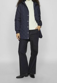 Granatowa pikowana kurtka z kapturem, zatrzaskami i elastycznymi mankietami, noszona na kremowym golfie i ciemnych dżinsach, w połączeniu z czarnymi butami.