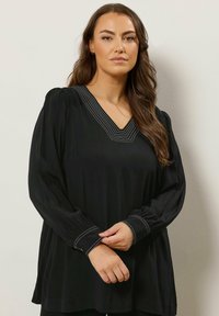 Blusa nera a maniche lunghe con scollo a V e cuciture bianche a contrasto lungo il collo e i polsini, caratterizzata da una vestibilità rilassata e una texture liscia.