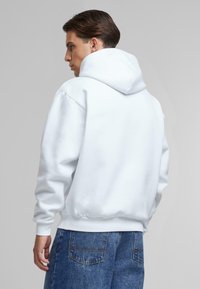 Haut à capuche blanc avec une grande capuche arrondie, des poignets côtelés et un ourlet côtelé. Présenté de dos, associé à un jean en denim bleu. Texture lisse.