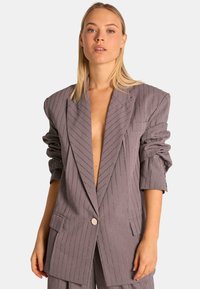 Blazer gris a rayas con hombros oversized, escote en V profundo, cierre de botones y puños enrollados. Presenta acentos de hilo metálico.