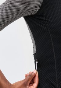 Χέρι που κρατάει το φερμουάρ στο πλαϊνό τσέπη ενός μαύρου υφασμάτινου μπουφάν με γκρι μανίκι με σχέδιο κυψελών.