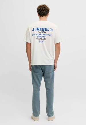 Homme aux cheveux bouclés portant un t-shirt blanc avec un motif graphique et un texte bleu, ainsi qu'un jean bleu clair, debout de dos devant un fond uni.