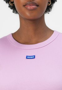 Paarse geribbelde shirt met een blauwe rechthoekige HUGO-logo patch op de borst. De halslijn is rond en glad van textuur.