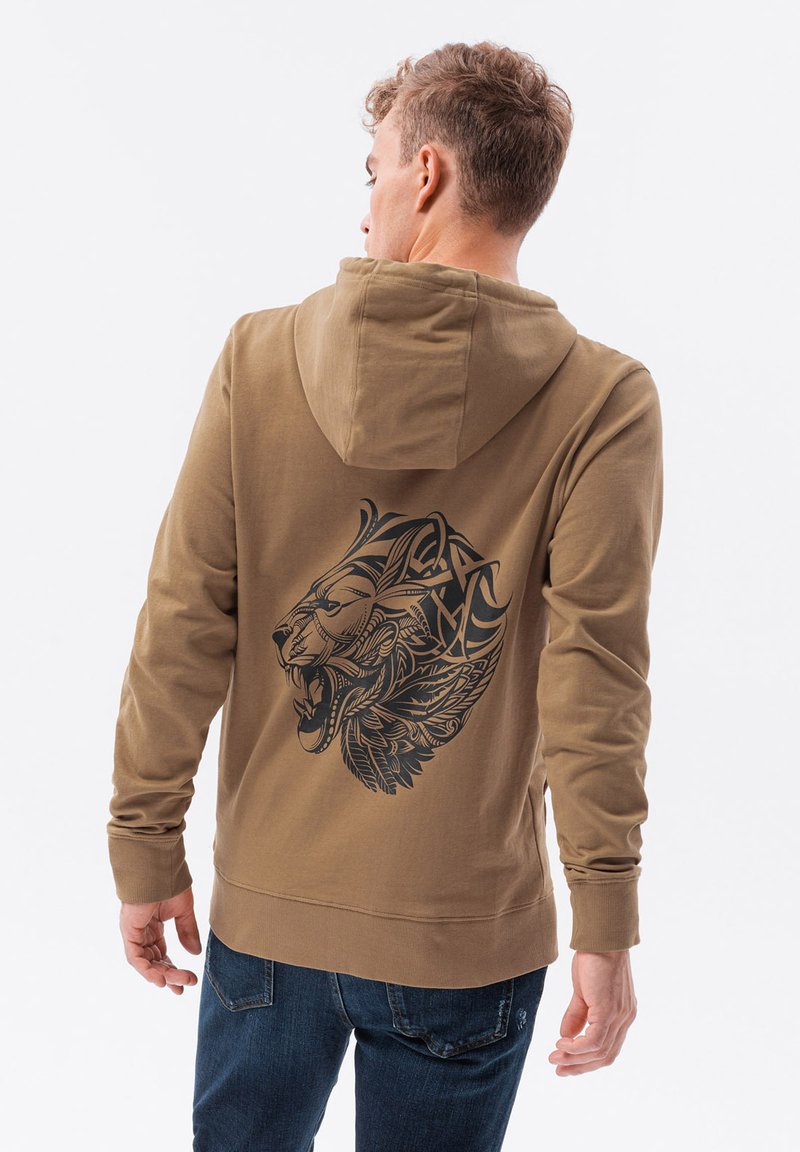 Felpa marrone con una grande grafica di un leone nero sul retro. Presenta un cappuccio con lacci e polsini a coste. Abbinata a jeans blu.