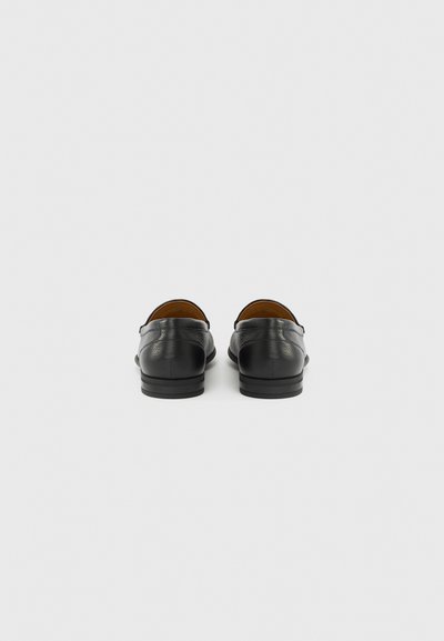 Ζευγάρι μαύρα δερμάτινα παπούτσια τύπου slip-on, εμφανιζόμενα από πίσω σε απλό λευκό φόντο.