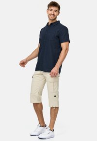 Camisa de botones de manga corta en navy con bolsillo en el pecho, combinada con pantalones cortos de cargo beige con bolsillos laterales y acabados con cordón. Zapatillas blancas.
