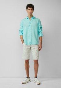 Camicia in lino azzurro chiaro con chiusura a bottoni, abbinata a pantaloni corti color crema e sneakers bianche con accenti scuri. Design semplice e casual.