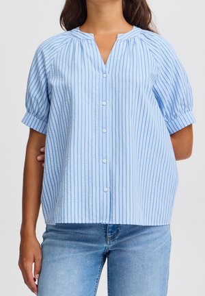 Femme portant un chemisier bleu clair à manches courtes, boutonné, avec des rayures verticales, et un jean bleu clair, posant devant un fond uni.