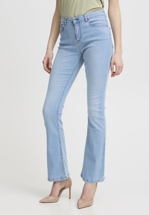 Kvinde iført lyseblå bootcut-jeans og beige højhælede sko, stående mod en ensartet hvid baggrund.