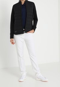 Doudoune sans manches noire avec coutures texturées, associée à un polo bleu marine, un pantalon blanc et des baskets blanches. Fermeture éclair et col montant.