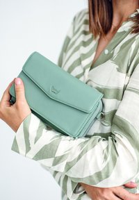 Mint green leather wallet with a flap closure and embossed logo, featuring a zippered side and smooth texture, held against a patterned shirt.