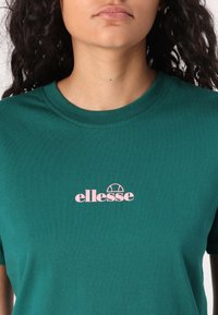 Mėlynas medvilnės marškinėlis su apvalia iškirpte, centre krūtinėje esantis rožinis "ellesse" logotipas. Lygus paviršius, trumpomis rankovėmis.