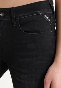 Jeans de mezclilla negra con un diseño ajustado, que presentan un bolsillo delantero, un botón plateado y una marca bordada en blanco en el bolsillo trasero.
