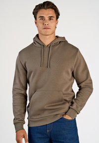 Sudadera con capucha marrón en tejido suave, con un bolsillo frontal tipo canguro y capucha con cordones. Combinada con jeans azules de corte clásico.