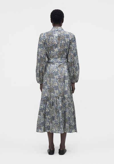 Άτομο που στέκεται με την πλάτη, φοράει μπλε φόρεμα με μακριά μανίκια και λουλούδια, με ζώνη και μαύρα παπούτσια σε λευκό φόντο.