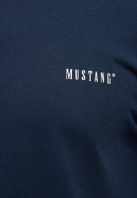 Námořnicky modrá látka s bílým velkými písmeny "MUSTANG®" potištěnými v oblasti hrudníku oděvu.