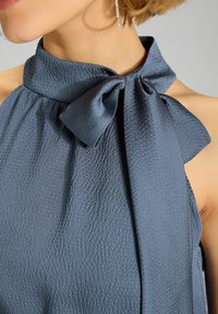 Navyblauwe jurk van een textuurstof, met een hoge kraag en een grote strikdetails bij de halslijn. Glad en gekreukt oppervlak.