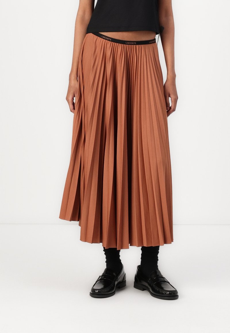 Femme portant une jupe midi plissée taille haute couleur rouille, avec des chaussettes noires, des mocassins noirs et un top court noir.