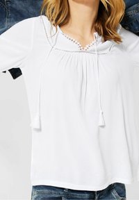 Vit blus med en texturerad yta, med rund halsringning, dekorativ spetskant och tofsar framtill. Matchas med denim.