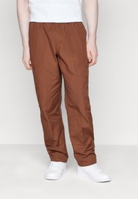 Man som har bruna cargobyxor med elastiskt midjeband och vita sneakers, som står mot en enkel ljus bakgrund.