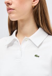 Γυναίκα που φοράει λευκό πόλο πουκάμισο με γιακά και πράσινο λογότυπο κροκόδειλου στο στήθος, καθώς και χρυσούς κρίκους στα αυτιά.