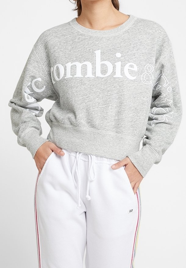 Sudadera gris claro con un logotipo en texto blanco en el pecho, con puños y dobladillo acanalados. Combinada con pantalones de chándal blancos con rayas laterales coloridas.
