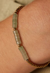 Kralenarmband met transparante groene cilindervormige kralen en glanzende goudkleurige bolvormige accenten. Draag je aan een pols met een lichte huidtextuur.