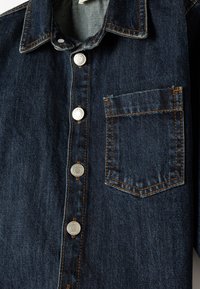 Camisa de mezclilla azul oscura con botones a presión de metal, un bolsillo en el pecho y detalles de costura naranja. Tejido texturizado con un diseño de cuello clásico.