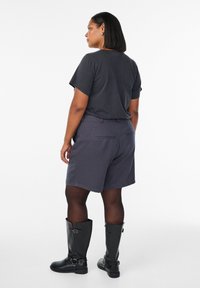 Kvinna med axellångt hår klädd i en svart t-shirt, mörkgrå shorts, genomskinliga svarta strumpbyxor och svarta knähöga stövlar, stående med ryggen mot oss.