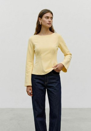 Kvinde iført en tætsiddende gul langærmet top og mørkeblå jeans, stående med den ene hånd nær taljen og kigger let mod højre.