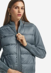 Leichte, gepolsterte Weste in sanftem Grau mit hohem Kragen. Mit Frontreißverschluss, seitlichen Taschen und gerippten, pulloverähnlichen Ärmeln.