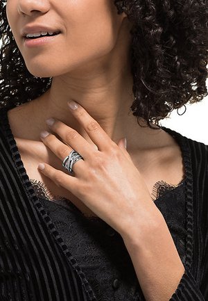 Silberner Ring mit ineinander verwobenen Designs, getragen an einer Hand mit hellem Nagellack. Der Hintergrund besteht aus einem schwarzen strukturierten Stoff.