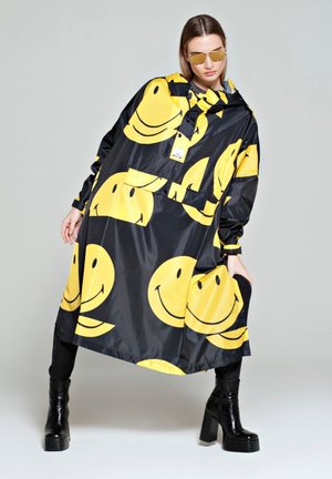Vrouw draagt een zwarte oversized regenjas met grote gele smileygezichten, zwarte plateau laarzen en geel getinte zonnebrillen tegen een effen achtergrond.